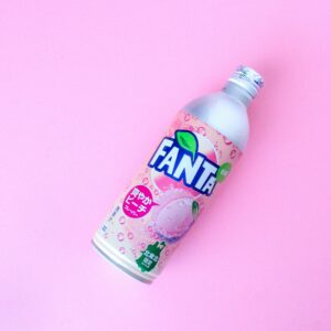 A vibrant peach flavored Fanta bottle displayed against a pastel pink background.