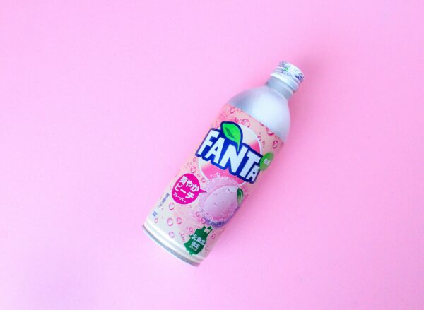 A vibrant peach flavored Fanta bottle displayed against a pastel pink background.